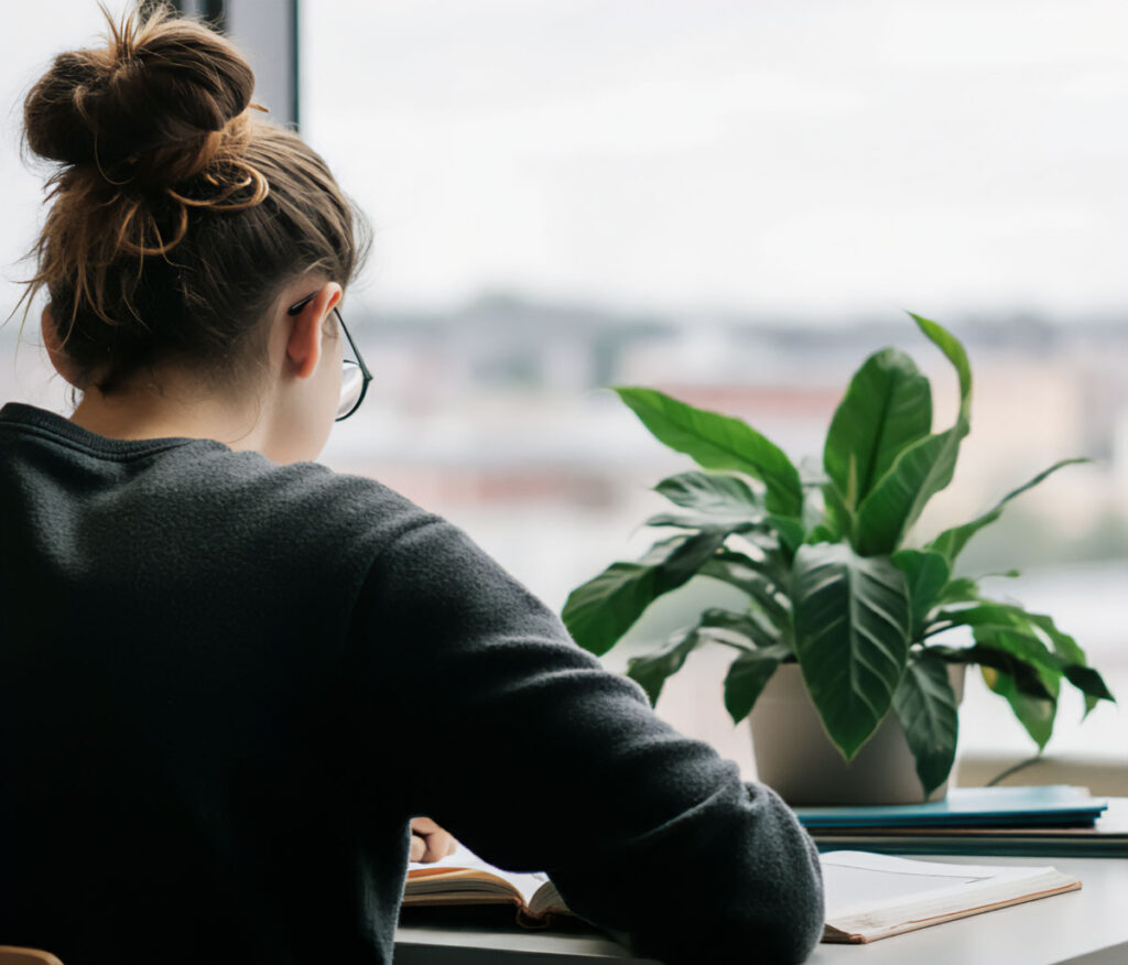 Woman reading by a window with a leafy plant on the desk beside her